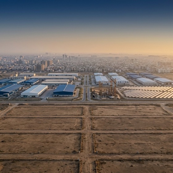 Aerial view at dusk of newly plotted State land beside warehouses and solar arrays on the edge of a Pakistani city